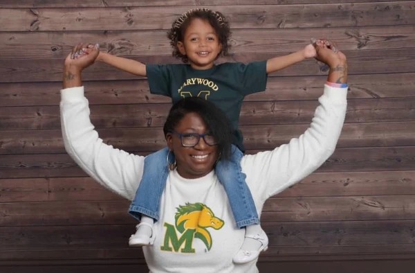 Smiling woman in a white Marywood University sweatshirt giving a piggyback ride to a young child in a matching t-shirt.